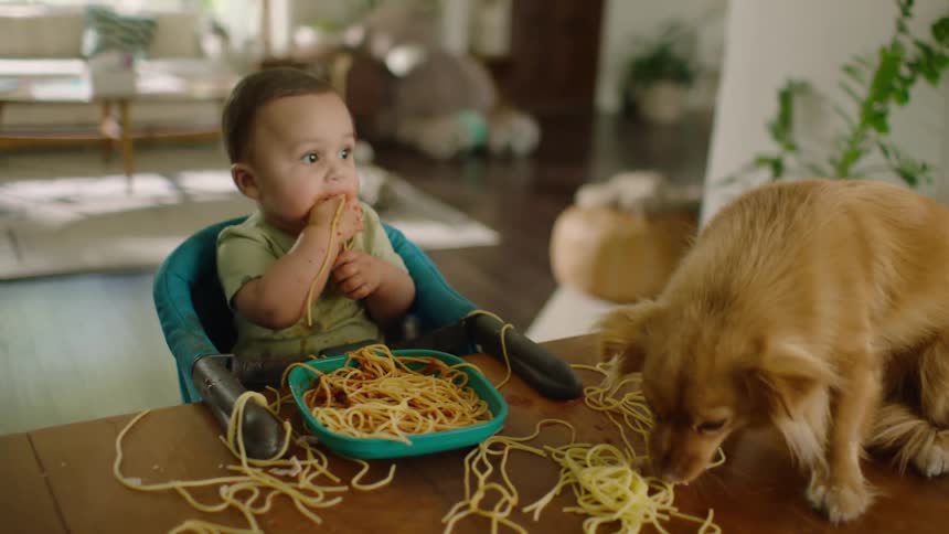 A funny scene of a baby and a dog sharing a messy spaghetti meal at the dining table