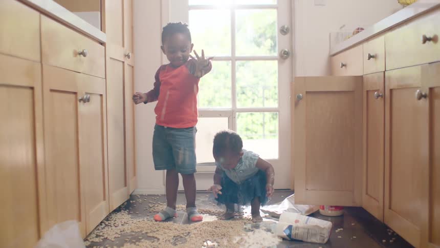 Mischievous young siblings gleefully play in a mess of cereal and flour they've spilled on the kitchen floor