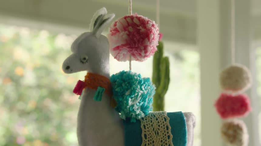 A cute handmade felt llama baby mobile with colorful pompoms gently sways in a sunlit room