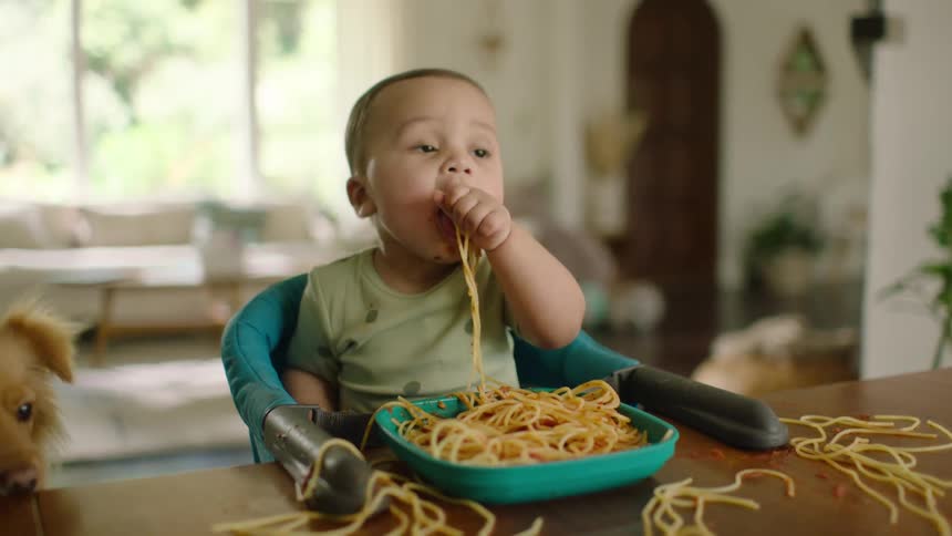 An adorable messy-faced baby in a booster seat at a table eats spaghetti with his hands