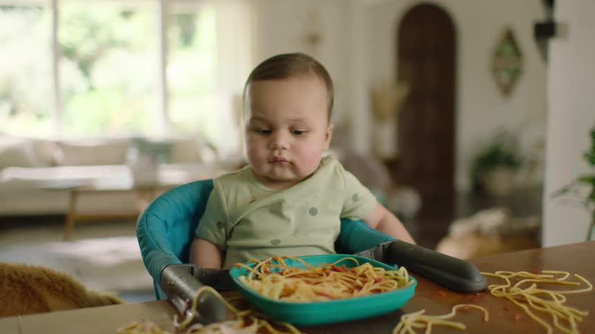 A cute baby watches their pet dog clean up the messy spaghetti on the dining table