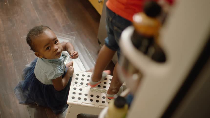 A high-angle view of a baby girl eating blueberries on the floor as her sibling stands on a step stool next to the refrigerator