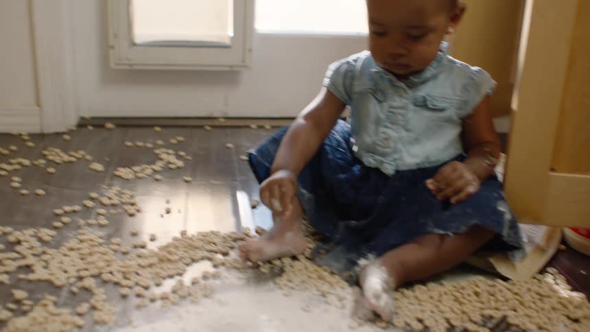 A young toddler girl sits on a wood floor playing in a big mess of spilled cereal and white powder in slow motion