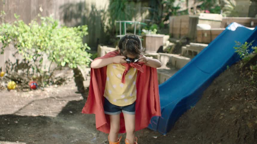 A young girl in a superhero costume with a red cape and goggles gets ready for an adventure in her muddy backyard
