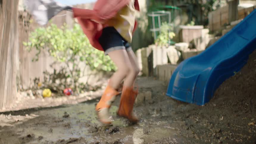 A young girl dressed as a superhero in a red cape and rain boots playfully jumps in a muddy puddle in slow motion
