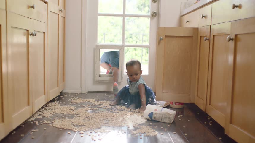 A baby girl plays in a mess of cereal and flour on the kitchen floor while her brother crawls out the doggy door