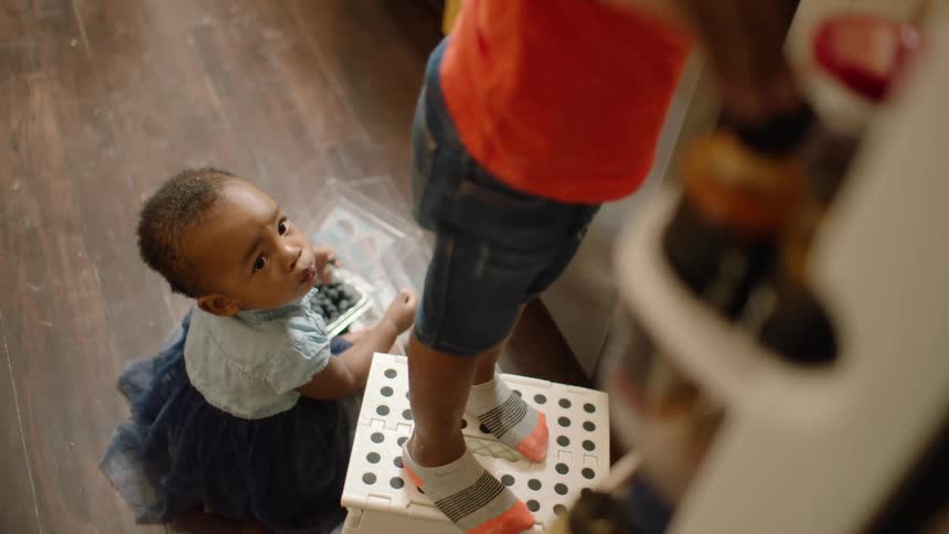 High-angle view of a toddler eating blueberries on the kitchen floor while their older sibling looks for a snack in the refrigerator