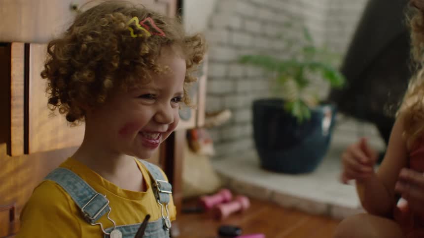 A happy little girl with curly hair and messy lipstick on her face smiles and laughs while playing with makeup
