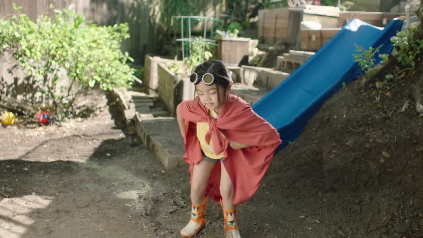 A little girl in a superhero costume with a red cape and rain boots joyfully stomps in a muddy puddle in slow motion