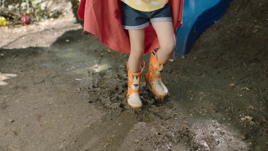 A slow-motion close-up of a young girl's rain boots stomping in mud reveals her superhero costume with a red cape and goggles as she plays in the backyard