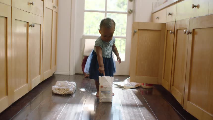 Cute baby girl stands innocently in the kitchen surrounded by a mess as her sibling crawls up behind her
