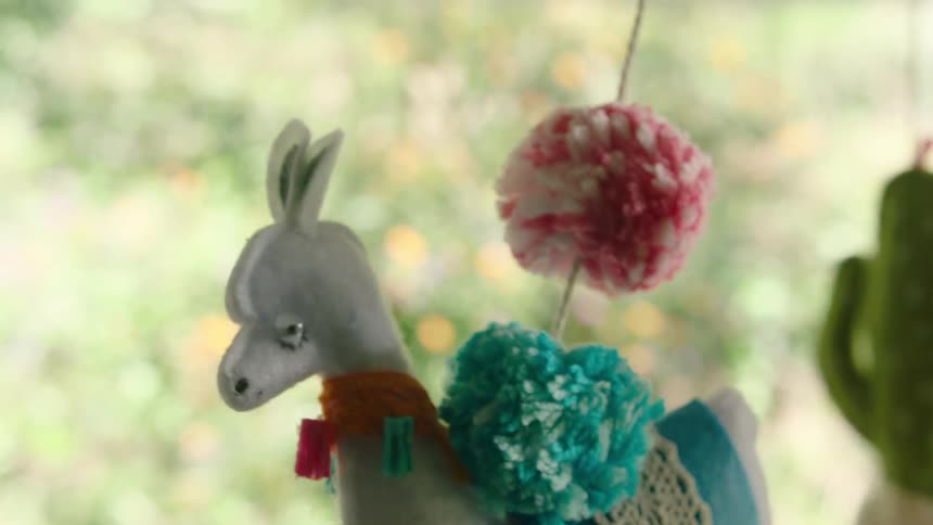 A colorful handmade felt llama ornament gently sways on a mobile in front of a window