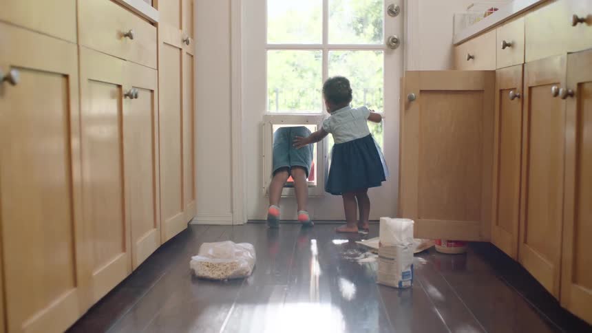 A young boy playfully climbs through a pet door to get outside as his toddler sister watches from the kitchen