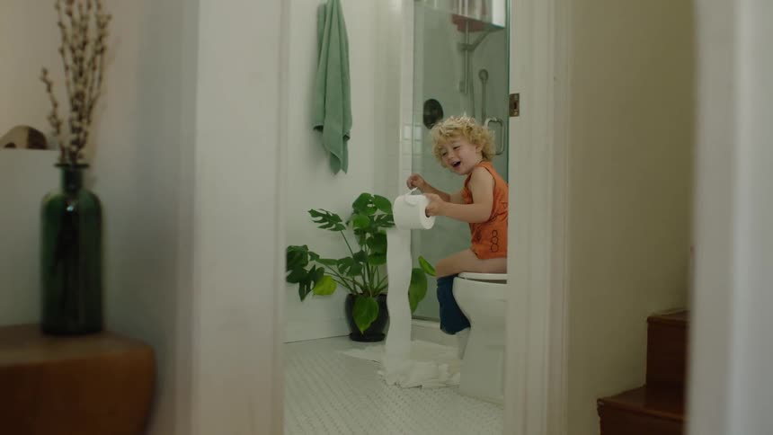 A cute, mischievous little boy laughs as he unravels a roll of toilet paper while sitting on the toilet in the bathroom