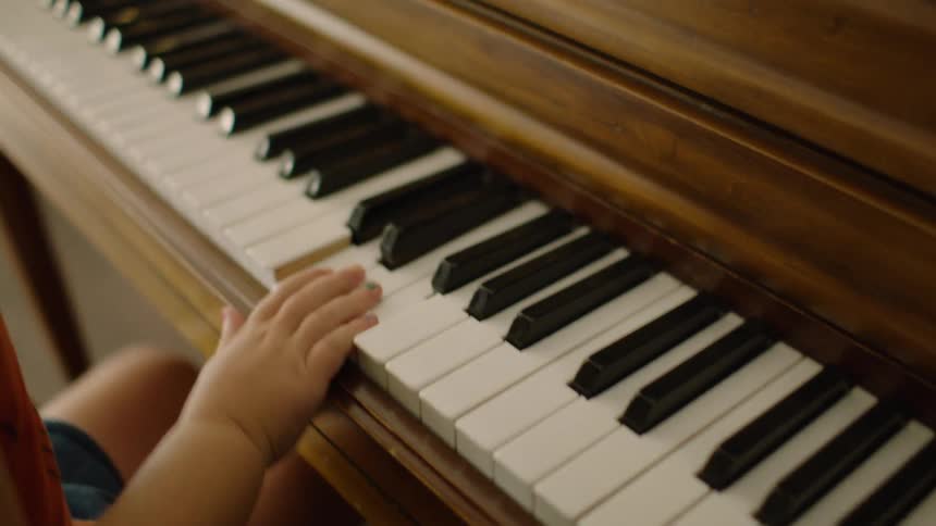 A young child's hands enthusiastically play on the keys of a classic wooden piano indoors