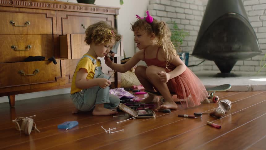 An older sister playfully puts makeup on her younger brother's face as they sit on the hardwood floor of their living room