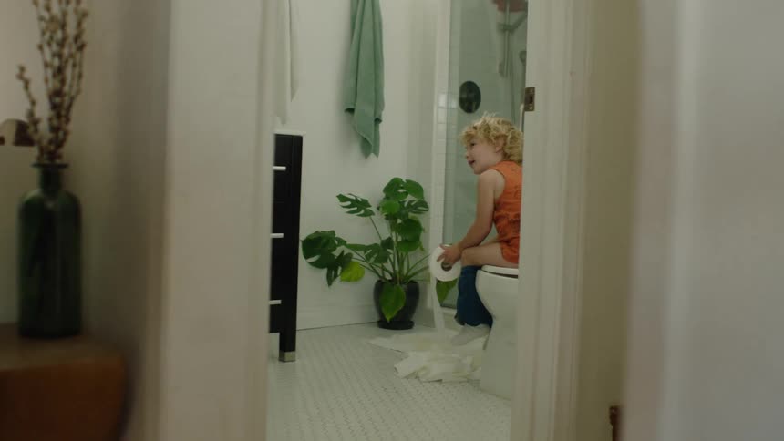 A mischievous little boy smiles at the camera while unrolling a long stream of toilet paper from the toilet in a stylish bathroom