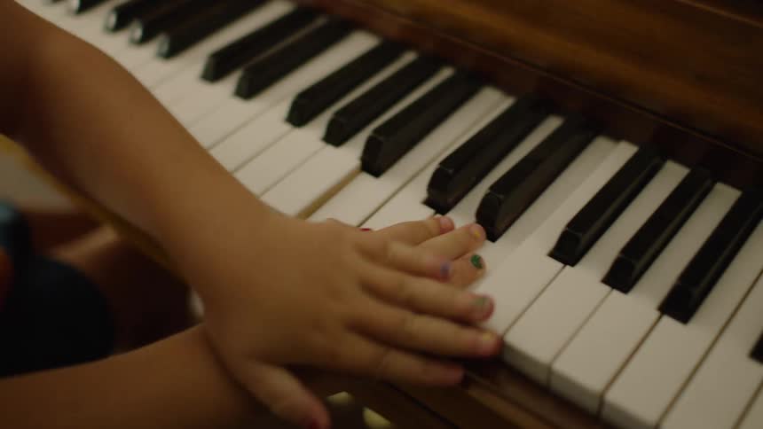 A close-up of a young child's hands with colorful painted nails playfully banging on the keys of a wooden piano