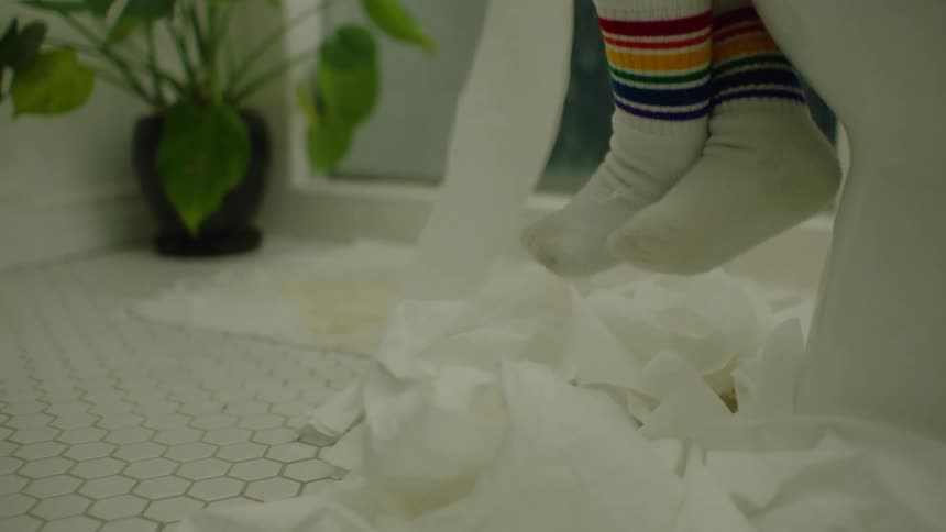 Close up in slow motion of a child's rainbow-socked feet swinging over a pile of unrolled toilet paper on the bathroom floor