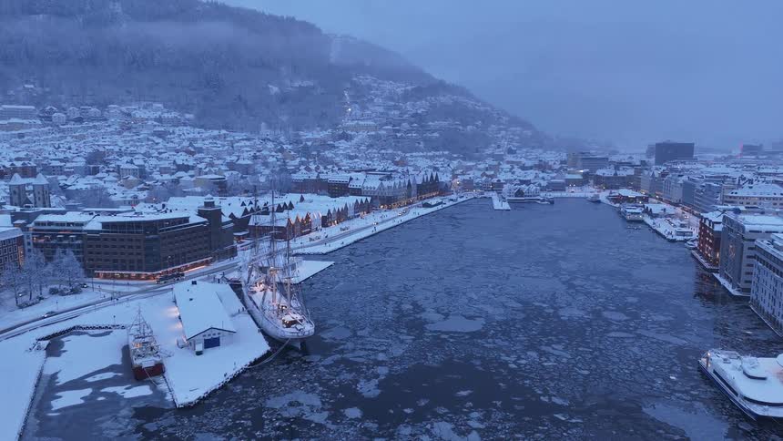 A serene aerial view of a snowy city harbor at dusk, where the water is filled with ice and a historic tall ship is moored by the snow-covered pier