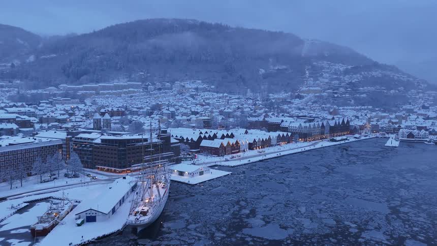 A scenic aerial view of the snow-covered Bryggen harbor in Bergen, Norway, featuring a tall ship docked in the icy fjord at twilight