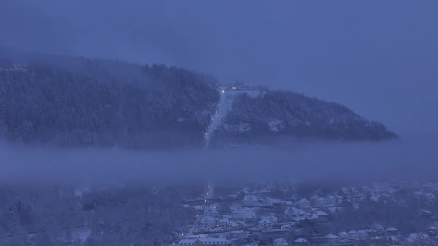A funicular ascends a snow-covered mountain above a town blanketed in thick fog during the quiet twilight hours