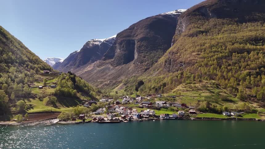 A scenic view reveals a tranquil village on the shore of a Norwegian fjord, surrounded by towering green and snow-capped mountains