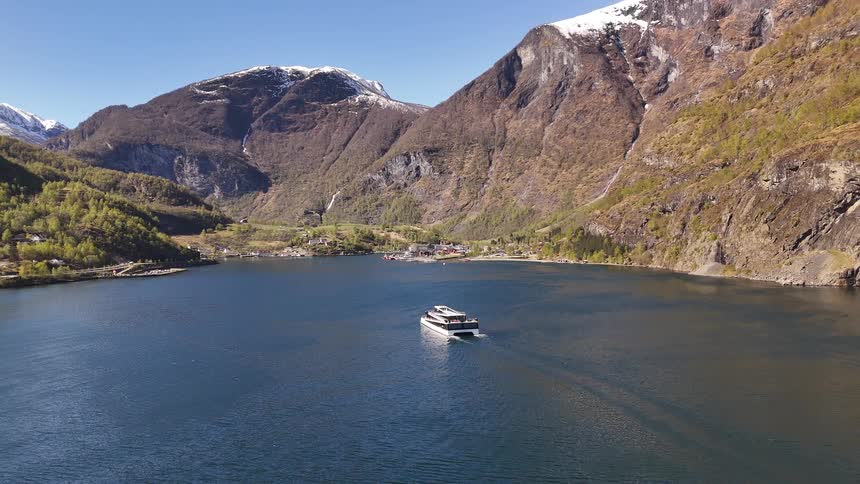 An aerial view shows a modern white ferry sailing across a calm fjord towards a remote village nestled beneath towering, snow-capped mountains in Norway