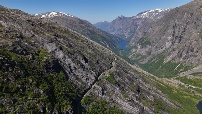 A stunning, static aerial view captures a narrow hiking trail etched into the side of a vast, rocky mountain range, with a deep valley and a serene lake below