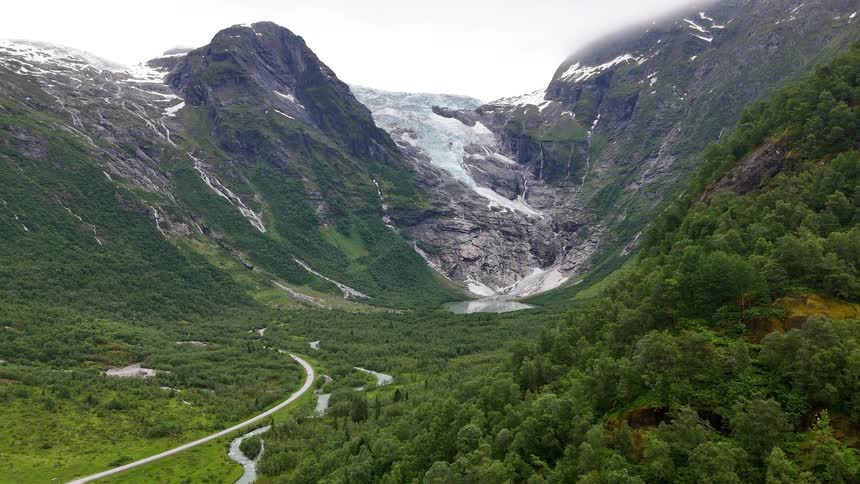 An aerial view reveals the majestic Boyabreen glacier and its meltwater river flowing through a lush green valley in Norway