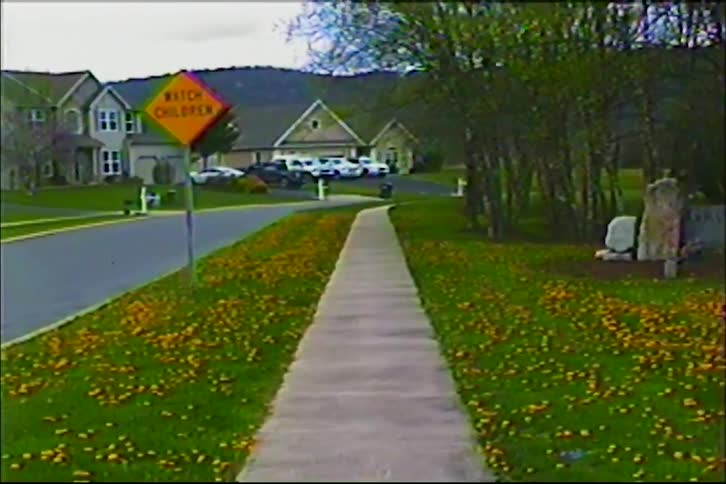 A vintage home video shot focuses on a quiet suburban sidewalk with a 'Watch Children' sign, dandelions in the grass, and houses in the background
