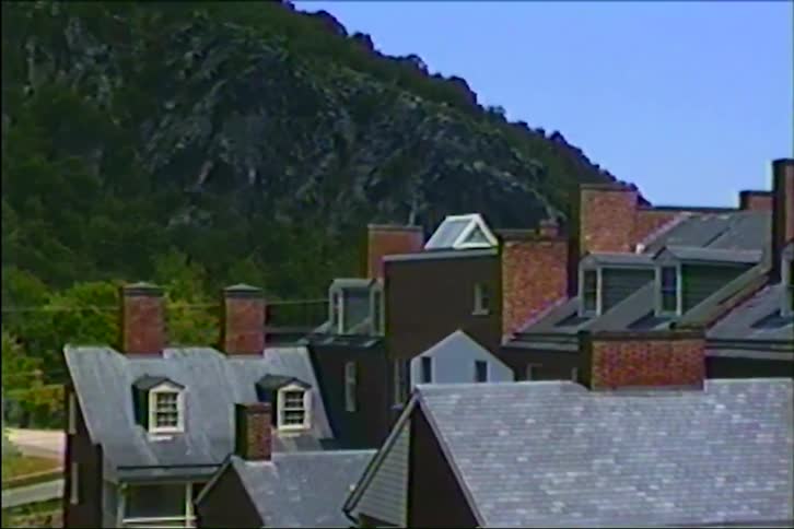 A slow zoom shot shows the brick chimneys and slate roofs of a town with a rocky, tree-covered mountain in the background