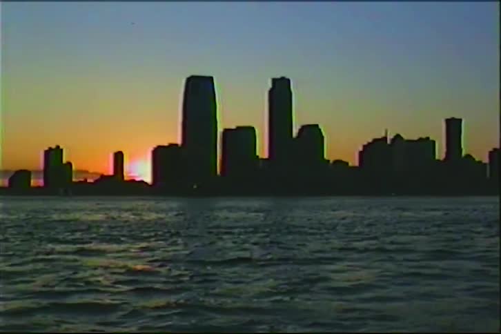 A vintage VHS shot shows the sun setting behind the silhouetted skyline of a city across the water