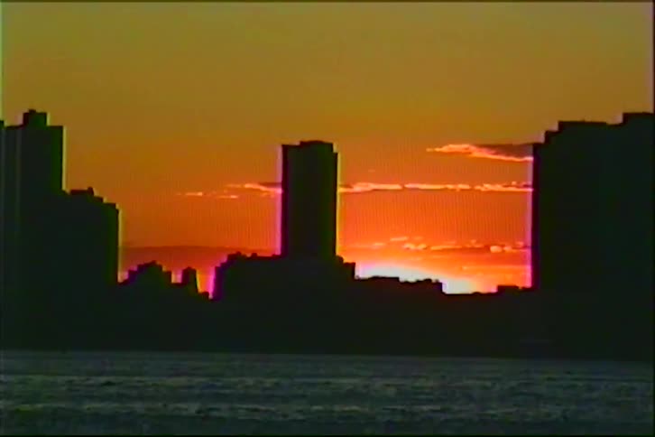 A vintage VHS shot shows the silhouette of a city skyline against a fiery orange sunset over the water