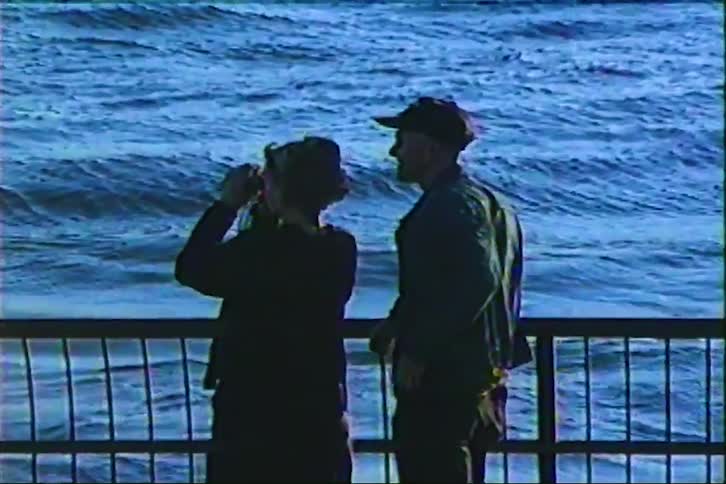 A couple shares a romantic kiss on a pier while looking out at the vast ocean through binoculars in this retro VHS footage