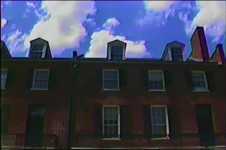Vintage footage shows a low-angle shot of historic red brick townhouses under a beautiful blue sky with white clouds