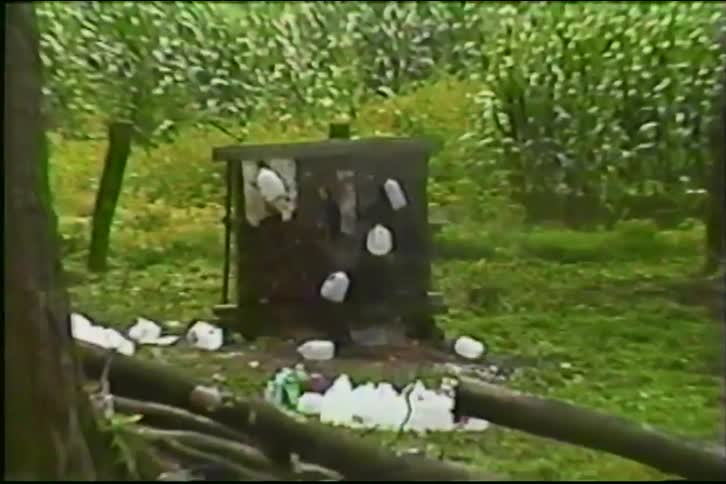 Vintage VHS footage slowly zooms in on a homemade shooting target decorated with plastic bottles in a wooded area next to a farm