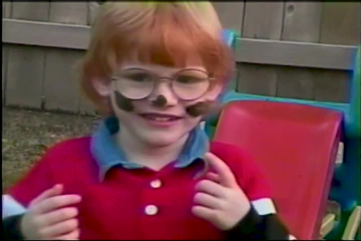 A cute, red-haired boy with glasses and his face painted like a puppy smiles for the camera in a nostalgic home video