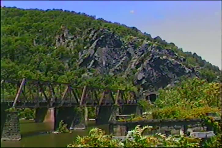 A vintage recording shows a static view of a railroad bridge crossing a river with a verdant, rocky mountain in the background