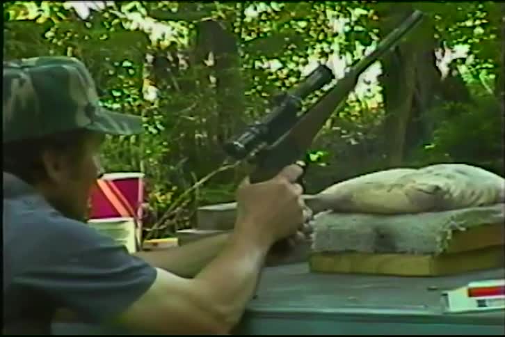 A man in a camouflage hat fires a pistol with a scope at a wooded outdoor shooting range in slow motion