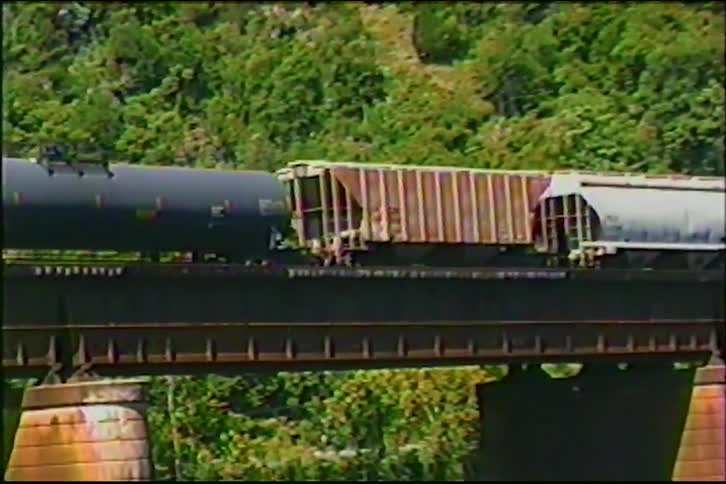 A freight train carrying various cargo cars crosses a scenic railroad bridge over a river at the base of a forested mountain
