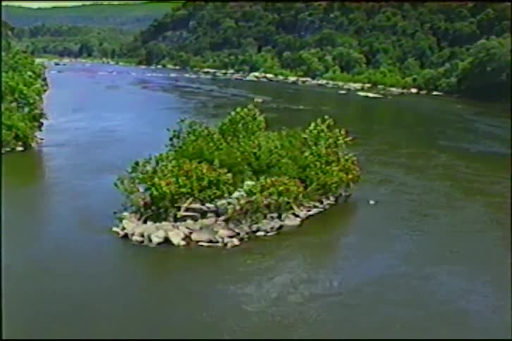 Vintage footage shows a slow zoom out over a tranquil river, featuring a small tree-covered island and dense green forested hills