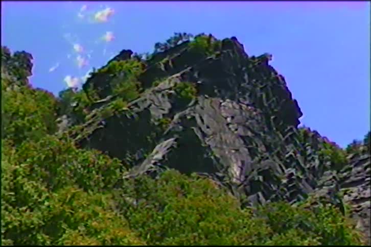 A low-angle shot slowly zooms in on a rugged, tree-covered mountain peak against a blue sky, captured on vintage VHS tape