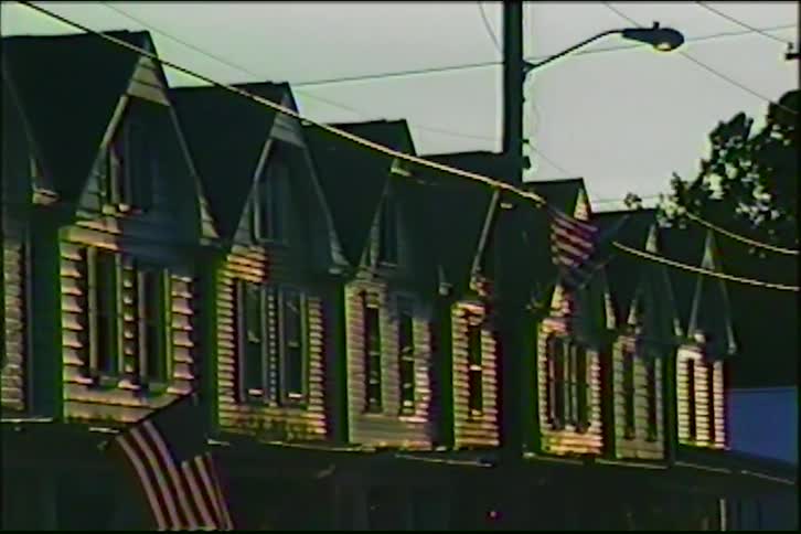 A slow pan across a row of traditional American townhouses with flags hanging outside, captured on vintage VHS film