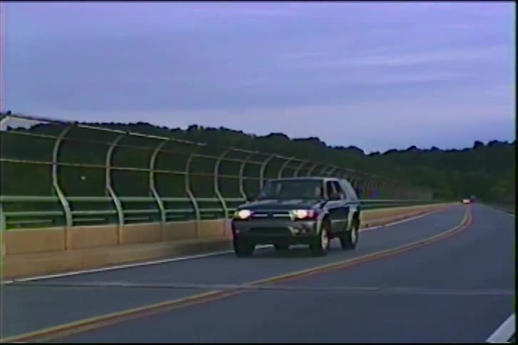 A dark green SUV with its headlights on drives towards the camera on a winding highway at dusk
