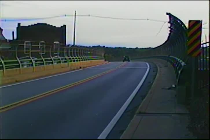 Vintage VHS footage captures a car and then a motorcycle driving across an empty urban overpass on an overcast day