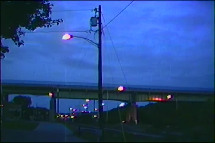 A vintage VHS-style shot of a street with glowing lights and an overpass as traffic passes in the distance at dusk