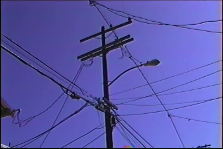 A chaotic tangle of power lines and cables crisscross around a telephone pole against a clear blue sky