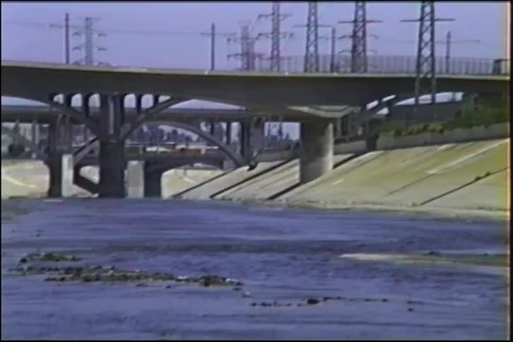 A dark bird flies in slow motion along a concrete urban drainage canal under industrial bridges