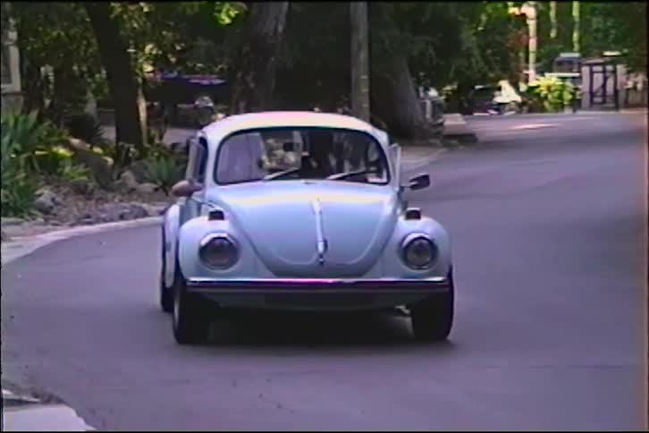 A classic blue Volkswagen Beetle drives toward the camera on a neighborhood street in this archival clip